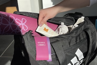 A hand holding a tube of muscle relief cream is reaching into a black sports bag. Inside the bag, there is a pink rolled-up yoga mat with floral patterns and a purple heavy resistance band. The bag also contains white items, possibly clothing.