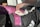 A hand holding a tube of muscle relief cream is reaching into a black sports bag. Inside the bag, there is a pink rolled-up yoga mat with floral patterns and a purple heavy resistance band. The bag also contains white items, possibly clothing.