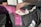 A hand holding a tube of muscle relief cream is reaching into a black sports bag. Inside the bag, there is a pink rolled-up yoga mat with floral patterns and a purple heavy resistance band. The bag also contains white items, possibly clothing.