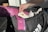 A hand holding a tube of muscle relief cream is reaching into a black sports bag. Inside the bag, there is a pink rolled-up yoga mat with floral patterns and a purple heavy resistance band. The bag also contains white items, possibly clothing.
