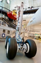 A large aircraft landing gear with intricate mechanical components and cables is displayed in a museum setting. Nearby is a helicopter with a navy insignia on it. Above, there appears to be a red object resembling a small buoy or vessel labeled 'DOUBLE EAGLE.' The background includes structural beams and museum displays.