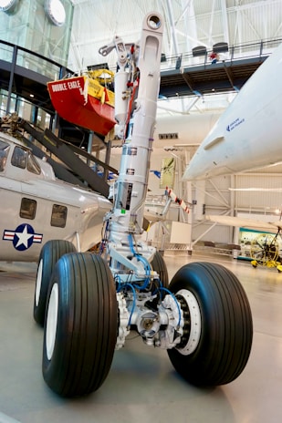 A large aircraft landing gear with intricate mechanical components and cables is displayed in a museum setting. Nearby is a helicopter with a navy insignia on it. Above, there appears to be a red object resembling a small buoy or vessel labeled 'DOUBLE EAGLE.' The background includes structural beams and museum displays.