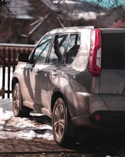 A silver SUV is parked outdoors with sunlight casting shadows of tree branches across its surface. The vehicle is positioned on a paved area with patches of snow around, indicating a winter setting. In the background, there is a wooden fence and a house with snow on the roof.