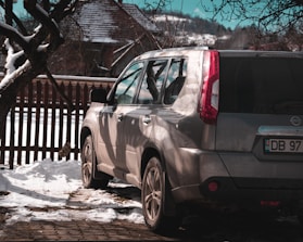 A silver SUV is parked on a snow-covered surface near a rustic wooden fence. The shadows of nearby trees fall across the vehicle, and a house with a snow-covered roof is visible in the background. Bare branches frame the upper part of the image, and the scene suggests a cold, wintry day.