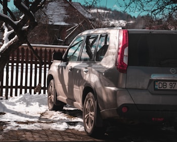 A silver SUV is parked on a snow-covered surface near a rustic wooden fence. The shadows of nearby trees fall across the vehicle, and a house with a snow-covered roof is visible in the background. Bare branches frame the upper part of the image, and the scene suggests a cold, wintry day.