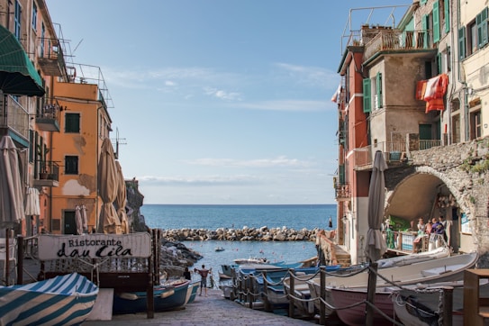The image depicts a picturesque coastal village with colorful buildings lining a narrow street leading to the sea. Small boats are docked along the sides, and people can be seen enjoying the scene. The architecture is charming, with shutters and balconies adding character to the buildings. A sign for a restaurant is visible, suggesting a lively and inviting atmosphere. The clear blue sky and calm sea contribute to a serene setting.