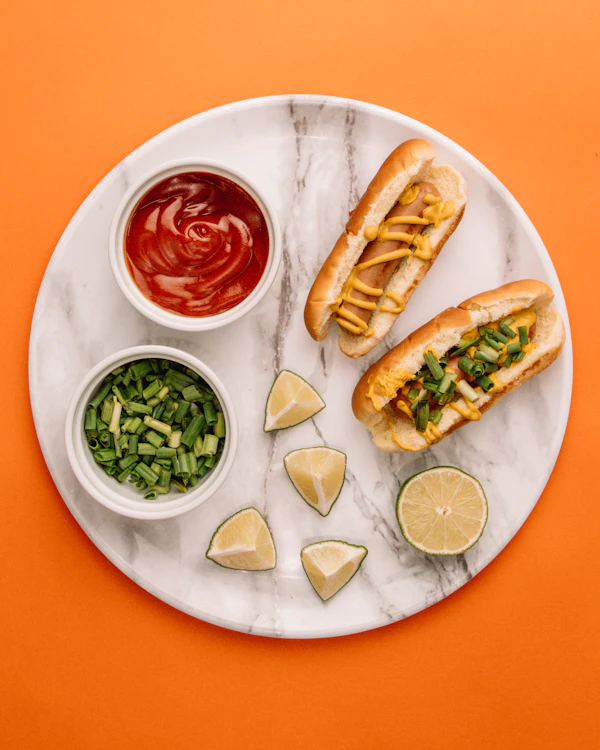 Two hot dogs on a marble board with dipping sauces and lime