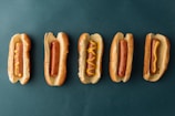 Five hotdogs in buns are arranged in a horizontal line against a dark green background. The hotdogs have various patterns of mustard on them, with the second one from the left having no mustard at all.