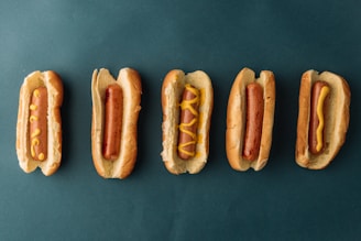 Five hotdogs in buns are arranged in a horizontal line against a dark green background. The hotdogs have various patterns of mustard on them, with the second one from the left having no mustard at all.
