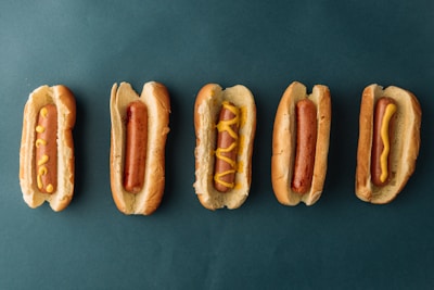Five hotdogs in buns are arranged in a horizontal line against a dark green background. The hotdogs have various patterns of mustard on them, with the second one from the left having no mustard at all.