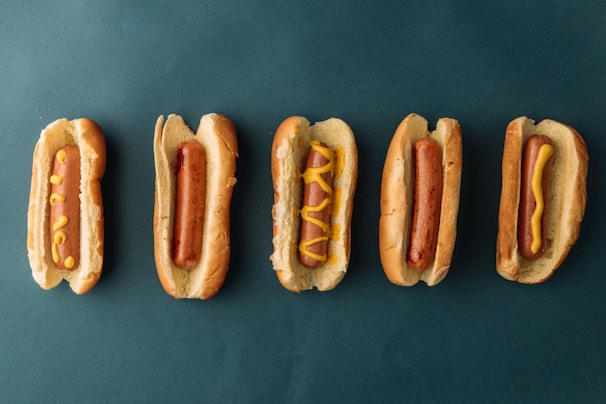 Five hotdogs in buns are arranged in a horizontal line against a dark green background. The hotdogs have various patterns of mustard on them, with the second one from the left having no mustard at all.