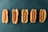 Five hotdogs in buns are arranged in a horizontal line against a dark green background. The hotdogs have various patterns of mustard on them, with the second one from the left having no mustard at all.