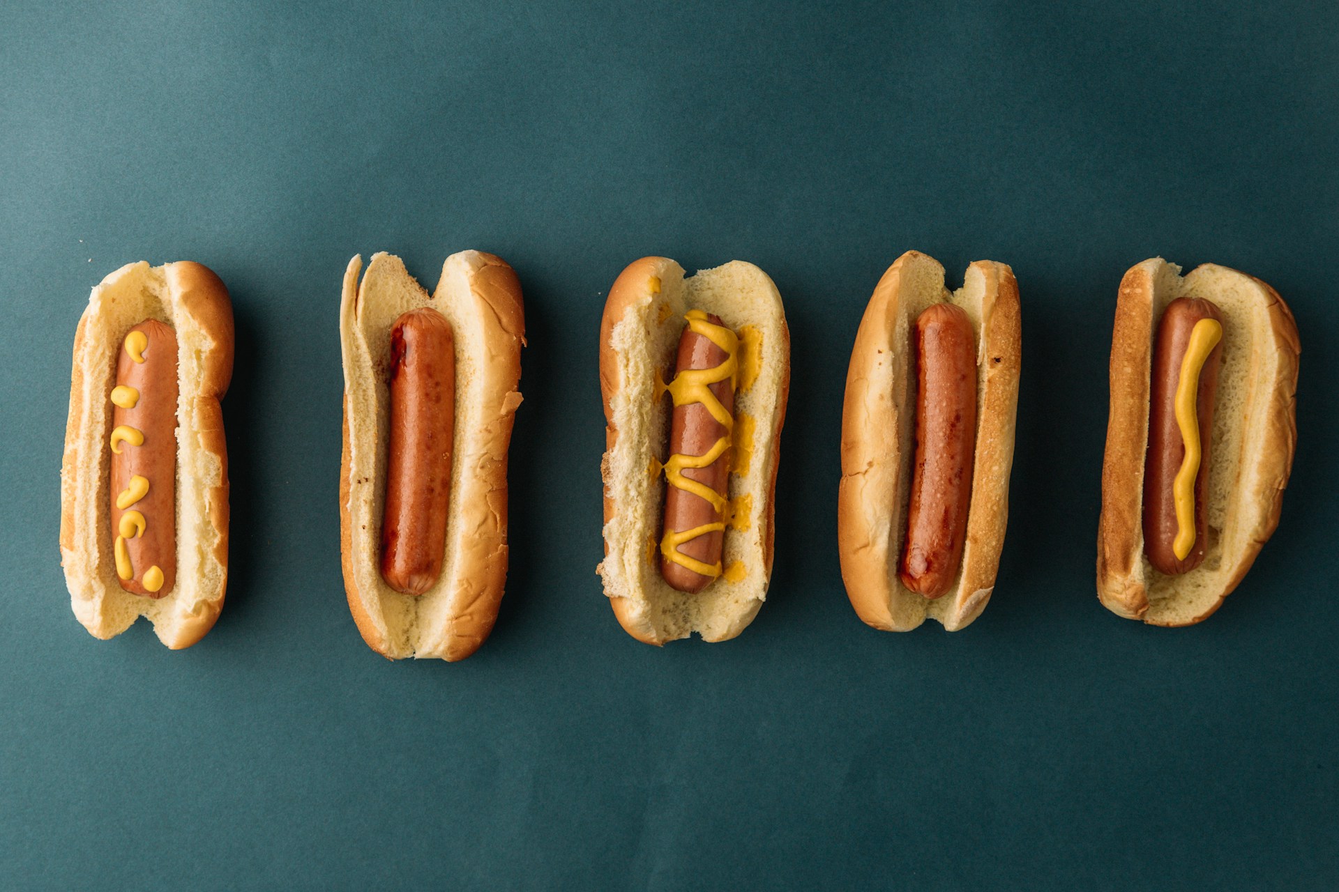 Five hotdogs in buns are arranged in a horizontal line against a dark green background. The hotdogs have various patterns of mustard on them, with the second one from the left having no mustard at all.