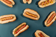 Several hot dogs are arranged on a dark teal background. Each hot dog is placed in a bun, with some topped with mustard and one including relish. The composition is neat and symmetric, with the hot dogs evenly spaced across the surface.