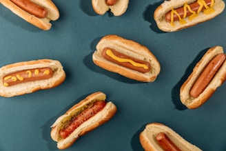 Several hot dogs are arranged on a dark teal background. Each hot dog is placed in a bun, with some topped with mustard and one including relish. The composition is neat and symmetric, with the hot dogs evenly spaced across the surface.