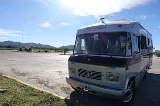 A vintage camper van with a distinctive design is parked in a sunny, open area. The van has a retro aesthetic with signs of wear, and its front is facing the viewer. The area surrounding the van is spacious with a paved surface and some greenery. In the background, there are mountains under a clear blue sky.
