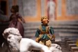 A marionette puppet with detailed features and clothing is positioned in the foreground. The puppet has a blue tunic with gold trims and beige pants. In the background, another puppet in a purple dress is partially visible. The setting appears to be indoors with a backdrop resembling a colorful, ornate architectural scene.