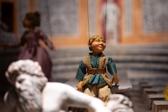 A marionette puppet with detailed features and clothing is positioned in the foreground. The puppet has a blue tunic with gold trims and beige pants. In the background, another puppet in a purple dress is partially visible. The setting appears to be indoors with a backdrop resembling a colorful, ornate architectural scene.