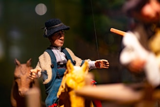 A detailed marionette puppet dressed in traditional clothing, including a hat and a textured coat, is positioned as if riding a carved wooden horse. The background is blurred, emphasizing the puppet in the foreground. The scene appears to be part of a larger diorama or display with other puppets or figures out of focus.