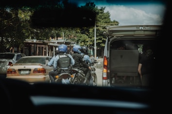 Three individuals wearing blue helmets and dark uniforms are seated on a motorcycle next to an open white van. They appear to be in an urban setting with cars parked nearby. Trees and buildings are visible in the background, suggesting a street scene.