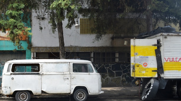 A white vintage van is parked beside a larger truck with a yellow and white paint scheme. The truck has spare tires attached to its side and writing on its rear panel. The setting includes a tree with overhanging branches and a building with a textured wall and a greenish facade partially visible behind the vehicles.