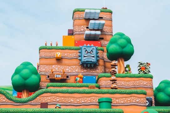 A colorful and vibrant landscape from a video game world featuring cartoon-like hills, animated trees, recognizable characters, and interactive elements such as blocks and clouds. The scene is layered with textures that resemble stone and grass, and includes elements like a large, menacing stone face and a stack of small creatures.