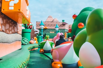 A vibrant theme park setting with colorful, large-scale models of video game elements. There are oversized features such as question blocks, turtle characters, and plant creatures. The scene is lively with cartoonish, exaggerated vegetation and structures, reminiscent of a popular video game universe.