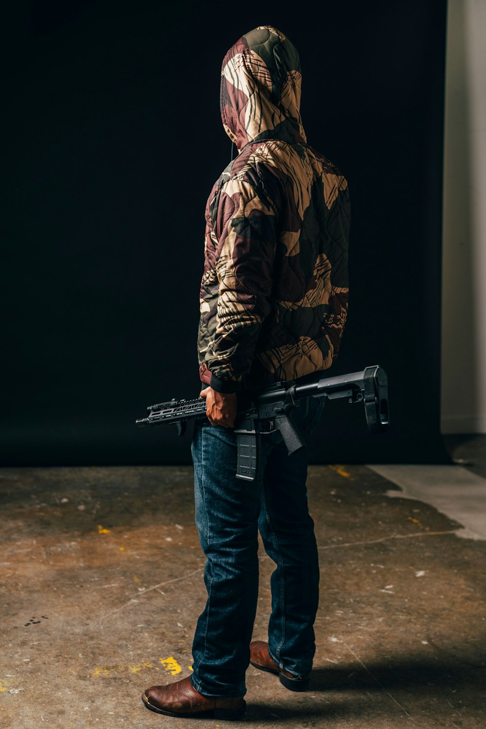 Hombre con chaqueta de camuflaje marrón y negro y jeans de mezclilla azules sosteniendo un rifle negro