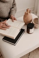 A person wearing a cozy knitted sweater is seated at a white table, reading an open book with another book underneath. Nearby are a clay pitcher, a candle in a jar, and a bowl containing twine.