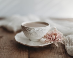 A delicate white teacup with an intricate pattern and saucer rests on a wooden surface. Beside the cup, a soft pink flower lies gently, adding a touch of elegance. The scene is softly lit, creating a serene and calming atmosphere.