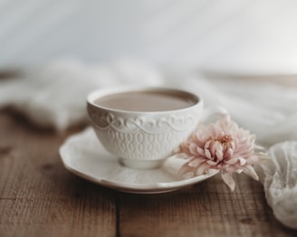 A delicate white teacup with an intricate pattern and saucer rests on a wooden surface. Beside the cup, a soft pink flower lies gently, adding a touch of elegance. The scene is softly lit, creating a serene and calming atmosphere.