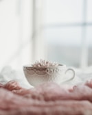 A beautifully ornate white teacup with intricate patterns sits on a soft, pink fabric surface. Inside the cup, pink flowers are arranged delicately. The background is softly blurred, giving a dreamy effect.