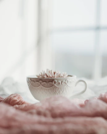 A pastel pink teacup adorned with delicate gold sparkles, sitting on a cream lace tablecloth with soft sunlight filtering through.