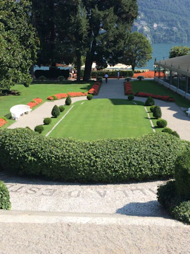 A beautifully manicured garden with a rectangular green lawn surrounded by neatly trimmed hedges and small topiary bushes. The garden leads to a scenic view of a lake, bordered by vibrant red flowers. Two people can be seen walking along a gravel path that runs alongside the lawn. A white bench is situated on the left, and there are trees and a building in the background.