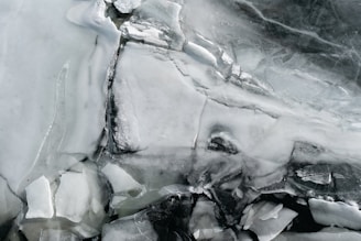 Translucent sheets and shards of ice form intricate patterns and textures, with variations in opacity creating a layered appearance. The surface is cracked and fragmented, revealing different shades of white, gray, and hints of darker tones.