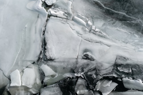 Translucent sheets and shards of ice form intricate patterns and textures, with variations in opacity creating a layered appearance. The surface is cracked and fragmented, revealing different shades of white, gray, and hints of darker tones.