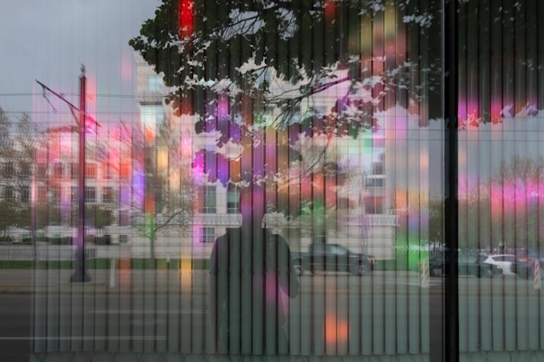 The image features a reflective glass surface that captures the silhouette of a person alongside an urban street scene. Behind the glass, there are vivid, colorful light patterns, possibly from an art installation, blending with the reflections of trees and surrounding buildings.