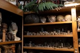 A wooden shelf filled with various objects, including ceramic and metal items, with some resembling traditional sculptures and carvings. There are several busts and masks to the left, and small metallic pieces organized neatly on shelves to the right. The setting appears like a workshop or studio with a mix of cultural artifacts and industrial components.