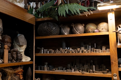 A wooden shelf filled with various objects, including ceramic and metal items, with some resembling traditional sculptures and carvings. There are several busts and masks to the left, and small metallic pieces organized neatly on shelves to the right. The setting appears like a workshop or studio with a mix of cultural artifacts and industrial components.