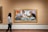 A person is walking in an art gallery, observing a painting. The painting features three individuals sitting at a table, engaging with printed materials. The vibrant artwork contains elements of expression and color, drawing attention to the subjects within a framed piece.