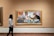 A person is walking in an art gallery, observing a painting. The painting features three individuals sitting at a table, engaging with printed materials. The vibrant artwork contains elements of expression and color, drawing attention to the subjects within a framed piece.