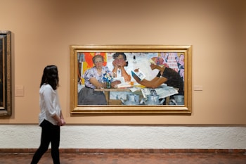 A person is walking in an art gallery, observing a painting. The painting features three individuals sitting at a table, engaging with printed materials. The vibrant artwork contains elements of expression and color, drawing attention to the subjects within a framed piece.