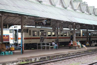 A train station platform with a train stopped beside it. Several people are sitting on the platform, some on benches and others on stools. There are signs hanging from the roof showing platform numbers 3, 7, and 6. The platform also has a vendor stall and a dog. The roof structure is supported by metal pillars and has a repetitive triangular pattern.
