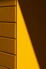 A wall with a geometric pattern of rectangular panels, half bathed in light and casting a sharp shadow. The light source creates a dramatic contrast between the illuminated and shaded areas, emphasizing the clean lines and texture of the panels.