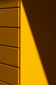 A wall with a geometric pattern of rectangular panels, half bathed in light and casting a sharp shadow. The light source creates a dramatic contrast between the illuminated and shaded areas, emphasizing the clean lines and texture of the panels.