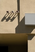 A textured concrete wall is illuminated by sunlight, creating defined shadows from a row of small insulators mounted on the wall. The shadows stretch diagonally across the surface, adding depth and contrast to the scene. A rectangular box, possibly a utility box, is attached to the wall.