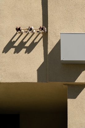 A textured concrete wall is illuminated by sunlight, creating defined shadows from a row of small insulators mounted on the wall. The shadows stretch diagonally across the surface, adding depth and contrast to the scene. A rectangular box, possibly a utility box, is attached to the wall.