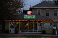 A florist shop with a prominently displayed neon sign reading 'Art Floral' in pink and green atop the entrance. The storefront windows display various plants and floral arrangements. The building has a quaint, cozy appearance, set against a backdrop of trees and a slightly overcast sky. Warm orange string lights are strung along the roofline, adding a welcoming touch.