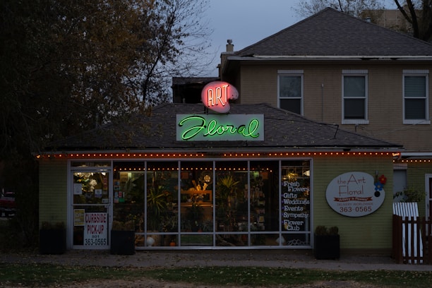 A florist shop with a prominently displayed neon sign reading 'Art Floral' in pink and green atop the entrance. The storefront windows display various plants and floral arrangements. The building has a quaint, cozy appearance, set against a backdrop of trees and a slightly overcast sky. Warm orange string lights are strung along the roofline, adding a welcoming touch.