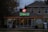 A florist shop with a prominently displayed neon sign reading 'Art Floral' in pink and green atop the entrance. The storefront windows display various plants and floral arrangements. The building has a quaint, cozy appearance, set against a backdrop of trees and a slightly overcast sky. Warm orange string lights are strung along the roofline, adding a welcoming touch.